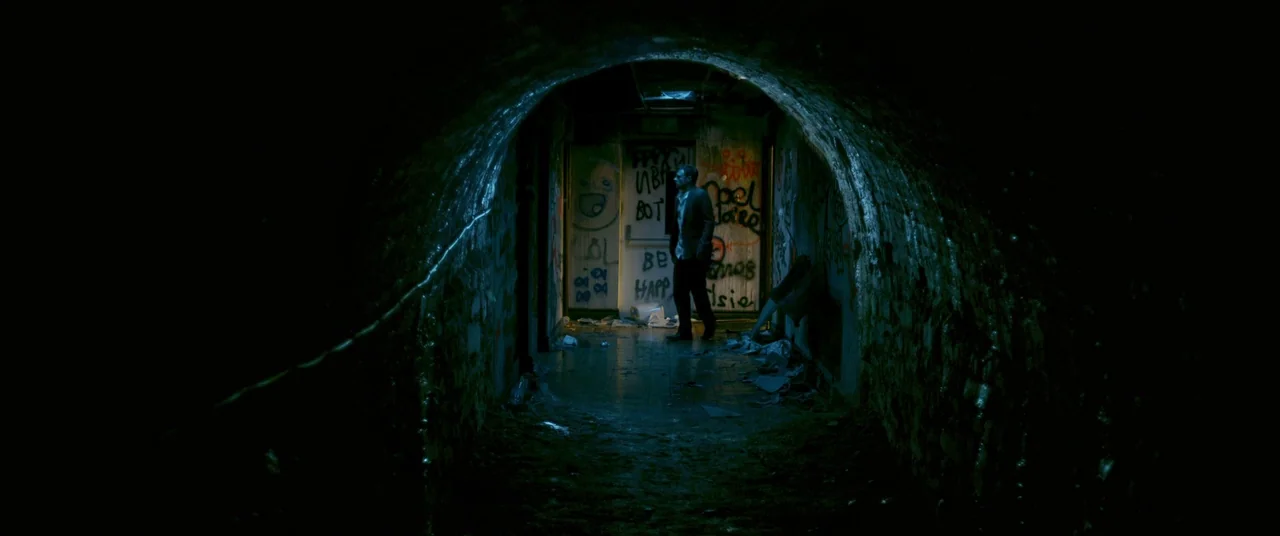 The Canal (2014) movie still: wide — An arched concrete tunnel features graffiti-covered walls and a single lit doorway at the end. A man stands at…