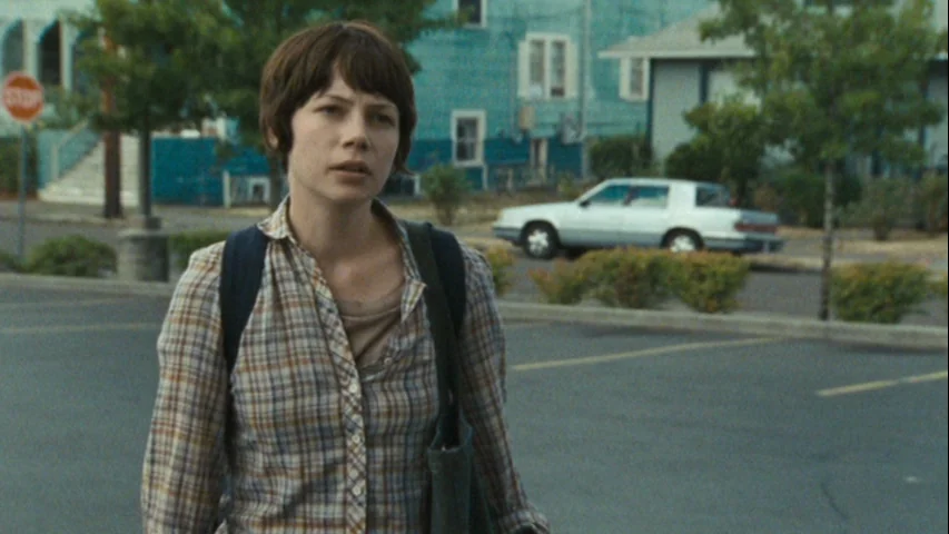 Wendy and Lucy (2008) movie still: medium — A woman walks through a sparsely occupied paved area near residential buildings. A turquoise house and a…