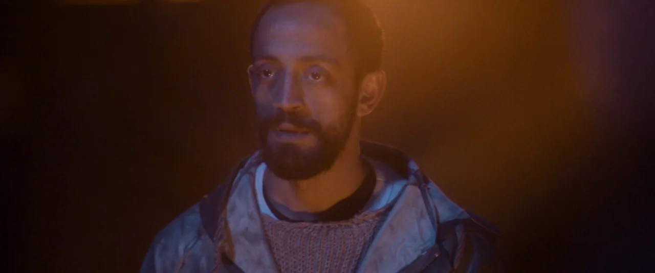 Baskin (2015) movie still: closeup — A man is illuminated by a warm glow in a dark, indistinct location. He turns his head and gazes toward the side…