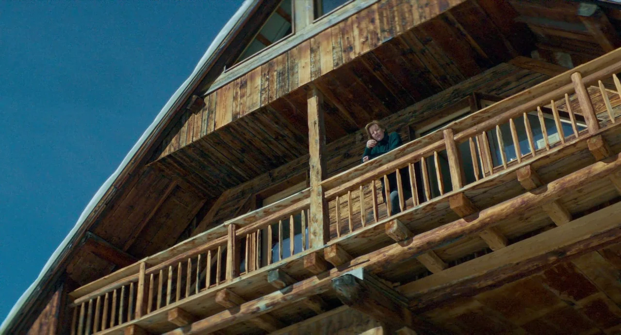 Anatomy of a Fall (2023) movie still: wide — A wooden balcony extends from a large cabin with visible roof rafters and siding. A woman in a dark…