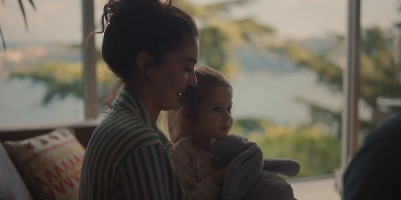 Kill Me If You Dare (2019) movie still: medium — A sunlit interior space with large glass windows overlooking greenery. A woman and child sit side…