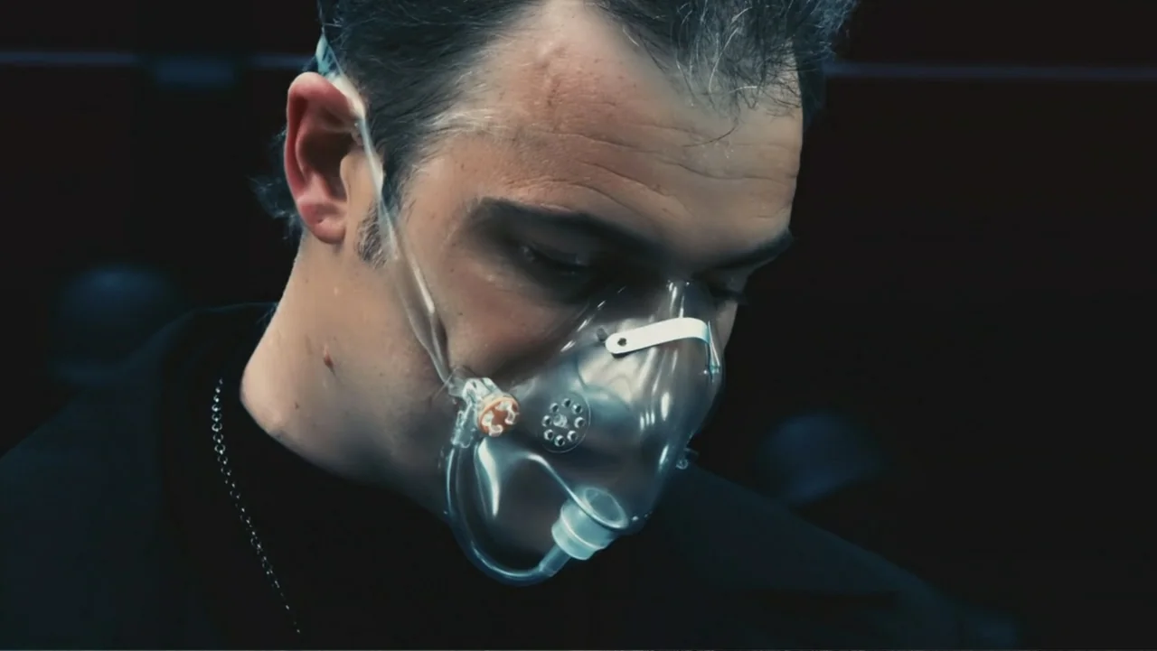 Ultraviolet (2006) movie still: closeup — A dark space with a man wearing a clear plastic oxygen mask. He tilts his head slightly while facing…