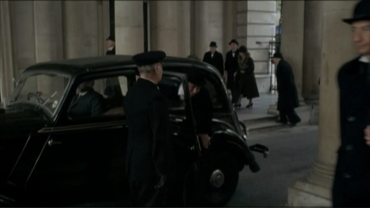 The Gathering Storm (2002) movie still: medium — A stone colonnade features tall pillars and a black vintage car parked at the curb. A chauffeur…