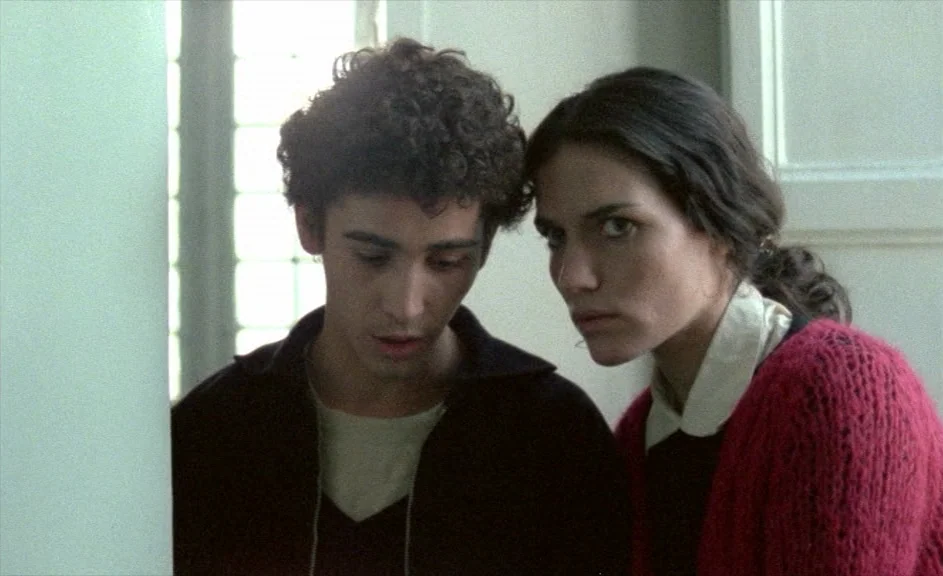 The Kingdom of Naples (1978) movie still: medium — Two young adults stand indoors against a plain light-colored wall with an adjacent window. The…