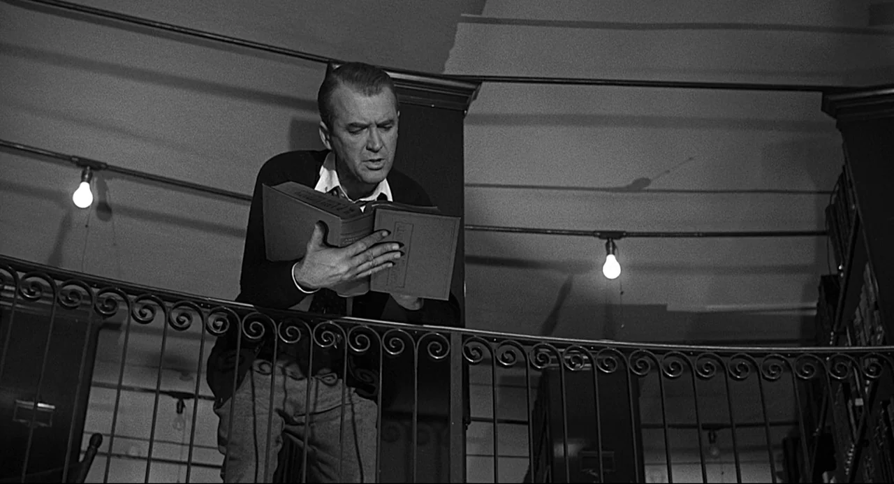 Anatomy of a Murder (1959) movie still: medium — A man stands on a metal railed mezzanine level inside a dark, quiet building. He leans over the…