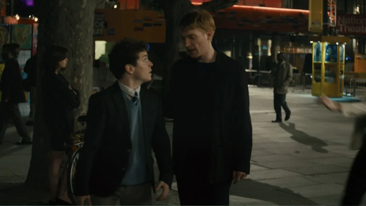 About Time (2013) movie still: medium — An urban outdoor plaza at night with paved ground and trees lining the path. Two men walk side-by-side…