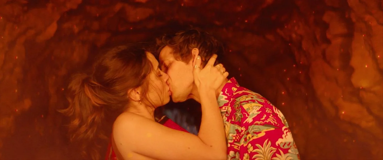 Palm Springs (2020) movie still: medium — A dark rocky cavern with red-tinged walls surrounds two people in a tight embrace. A man in a floral shirt…