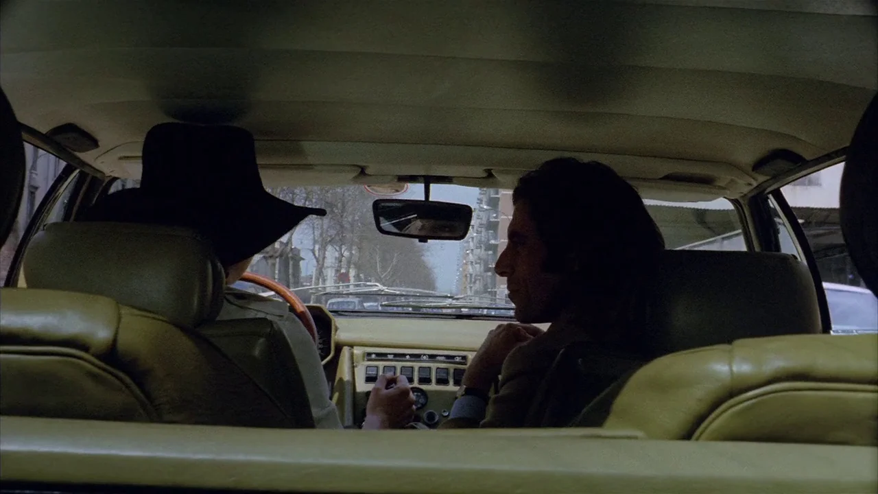Death Occurred Last Night (1970) movie still: medium — The interior of a 1970s car shows the dashboard and two front passengers. A driver wears a…