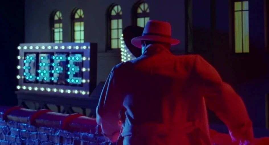 Dick Tracy (1990) movie still: medium — A dark urban rooftop features a glowing blue lightbulb sign that reads CAFE. A man wearing a fedora and…
