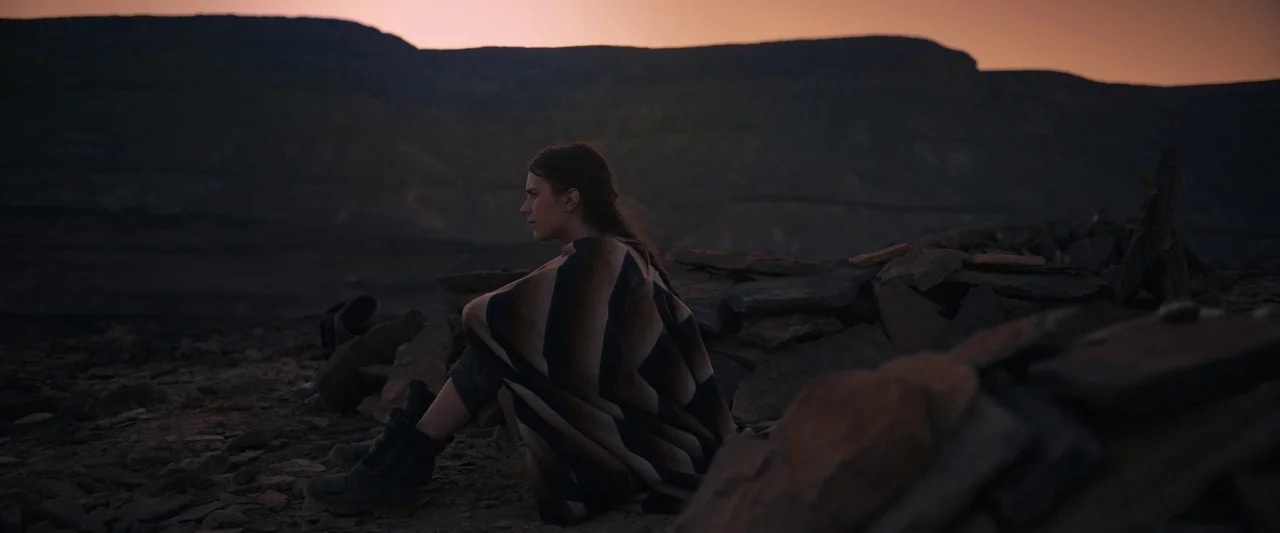 Settlers (2021) movie still: wide — A vast, desolate rocky terrain sits beneath a darkening sky at sunset. A young woman sits alone among scattered…