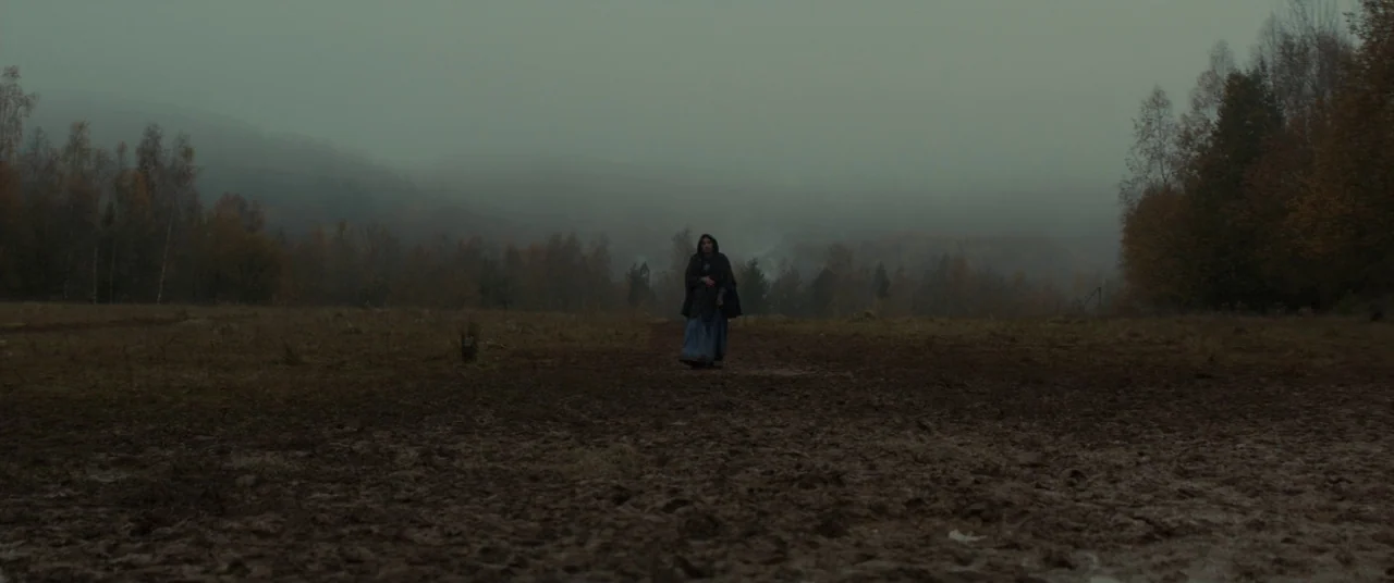Never Grow Old (2019) movie still: wide — A vast, muddy field stretches toward a treeline shrouded in thick morning fog. A lone woman in a hooded…