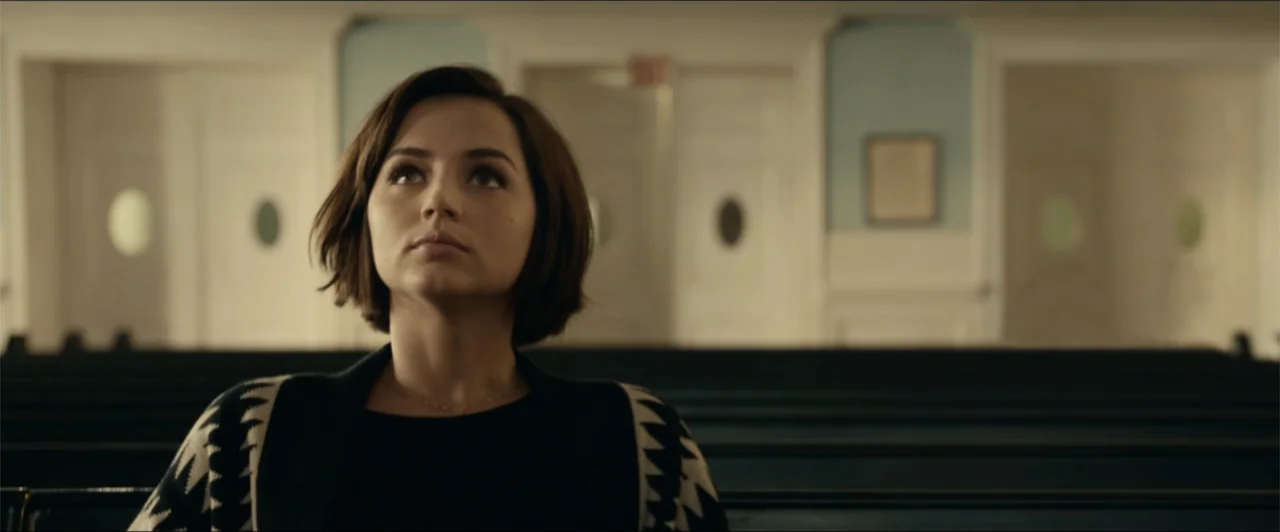 Exposed (2016) movie still: medium — A quiet church interior with rows of wooden pews and white doors in back. A young woman sits in a pew looking…