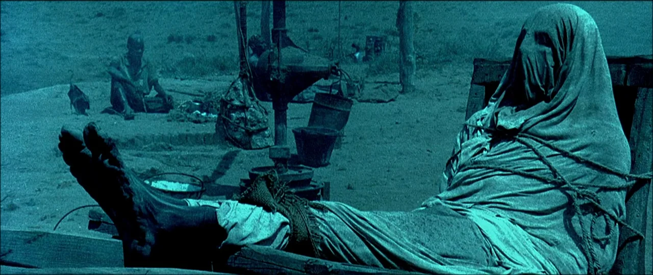 Black Hawk Down (2001) movie still: medium — A dusty desert location features a wooden structure and scattered metal buckets. A person is bound in…