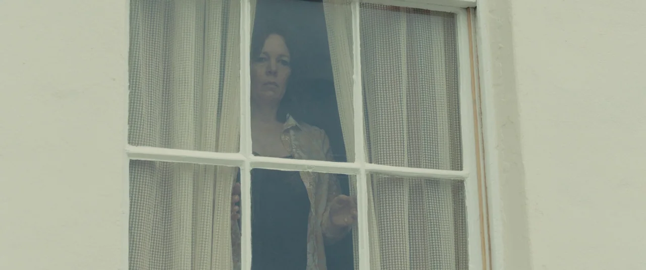 Empire of Light (2022) movie still: medium — A woman stands behind a multi-pane glass window with white frames. She holds back patterned beige…