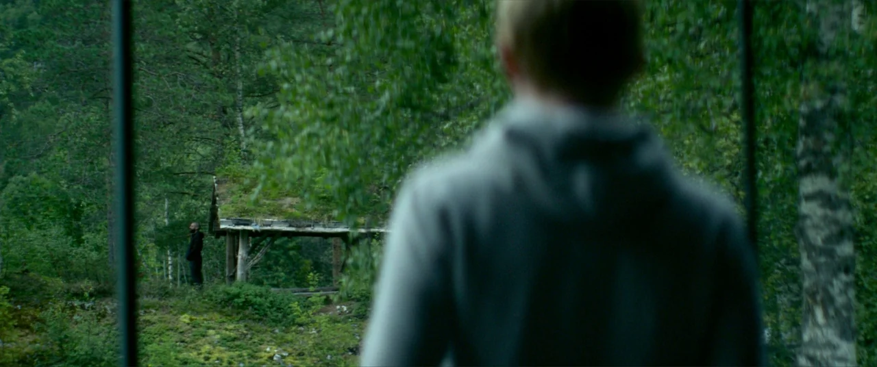 Ex Machina (2015) movie still: medium — A dense forest setting surrounds a rustic wooden structure with a grass roof. A person in a grey hood…