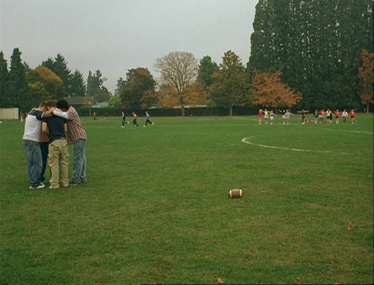 Elephant (2003) movie still: wide — An open grassy field with trees in the background during an overcast day. Three male teenagers stand in a tight…