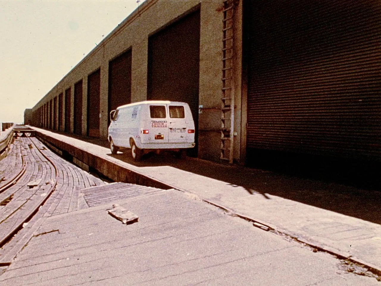 Space is the Place (1974) movie still: wide — A long industrial building with multiple metal roll-up doors sits on a concrete wharf. A white van is…