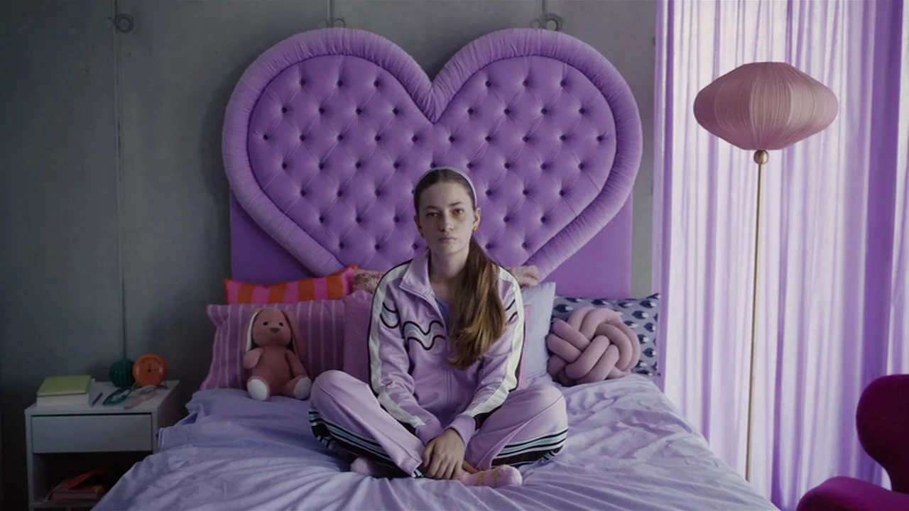 Club Zero (2023) movie still: medium — A modern room features a large heart-shaped purple headboard against a grey concrete wall. A young woman in a…