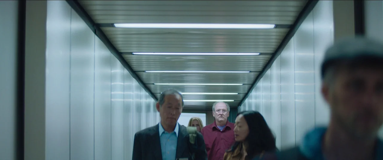 Kajillionaire (2020) movie still: medium — A sterile airport jet bridge with white panelled walls and overhead fluorescent lighting strips. Several…