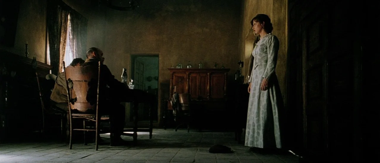 Renegade (2004) movie still: wide — A dimly lit room with stone floors and rustic furniture from the nineteenth century. A woman in a light dress…