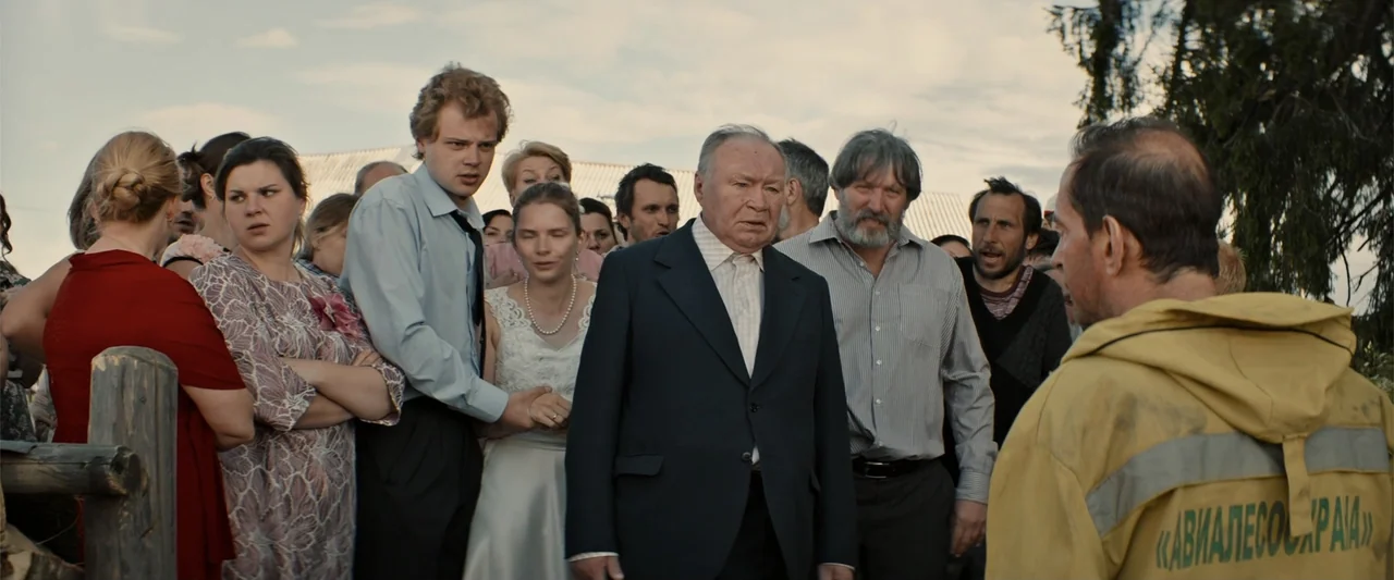 In the Line of Duty: Smoke Jumpers (1996) movie still: medium — An outdoor gathering of people in casual and formal attire on a cloudy day. An older…