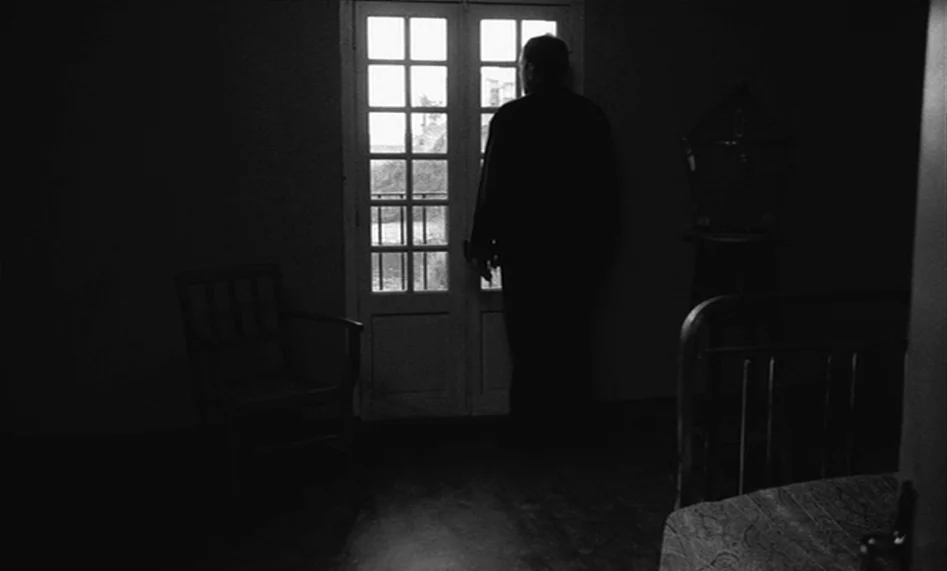The Man From London (2007) movie still: fullbody — A sparsely furnished, dark room features a glass-paned door looking out toward greenery. A man…