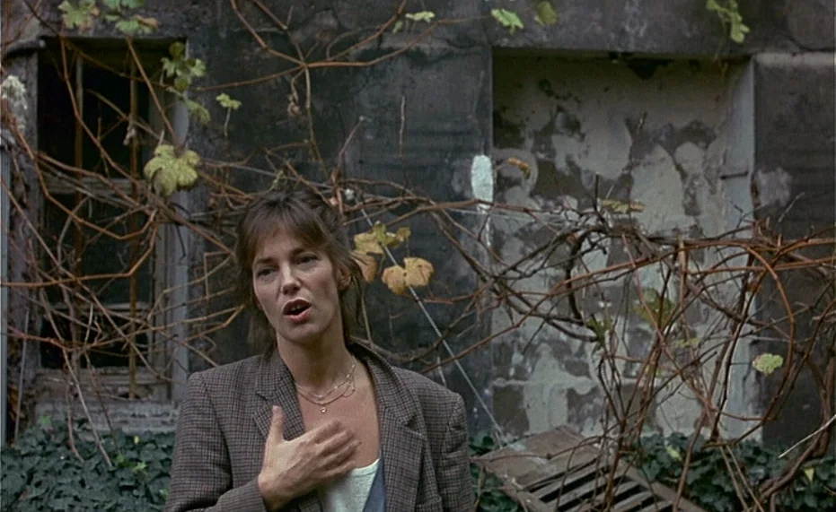 Jane B. par Agnès V. (1988) movie still: medium — A woman stands before a weathered brick wall covered in dried, leafless climbing vines. She keeps…