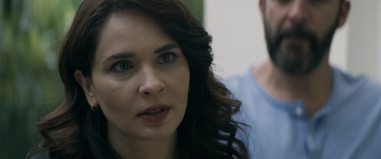 Human Animals (2020) movie still: closeup — A woman stands in the foreground with a man blurred behind her outdoors. They are engaged in a…