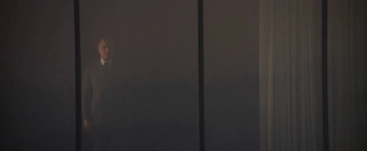 Klute (1971) movie still: wide — A man stands behind a grid of large reflective window panes in a building. The man remains perfectly still while…