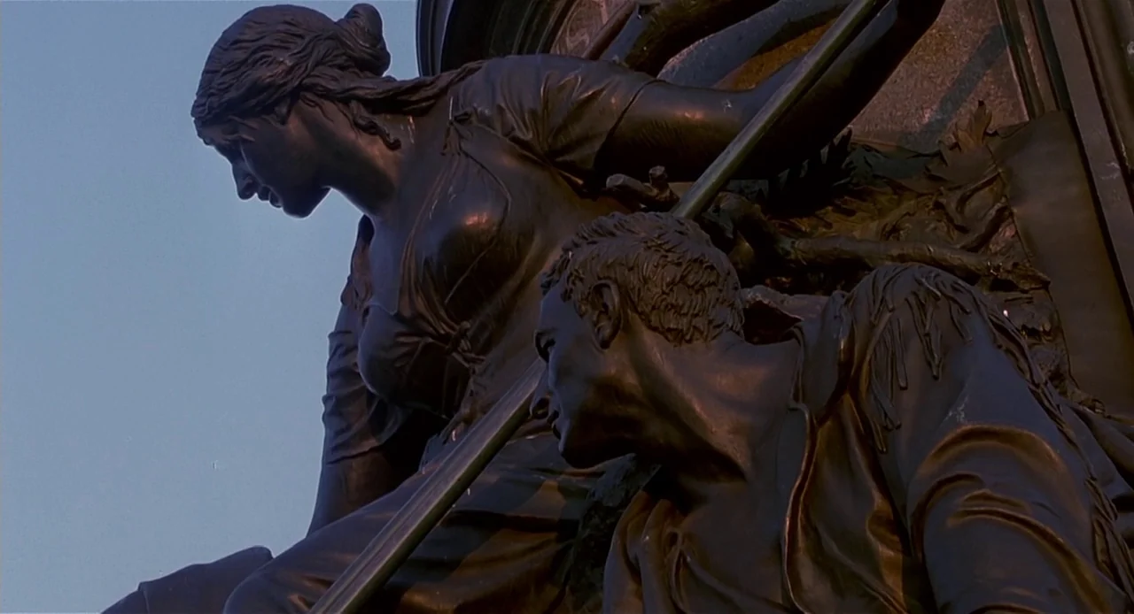 The Sixth Sense (1999) movie still: medium — A bronze memorial statue of a man and woman stands against a clear sky. The male figure looks upward…