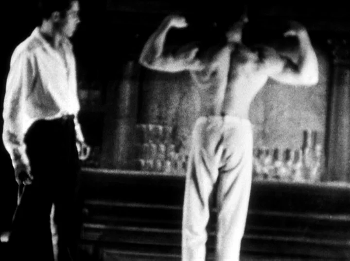 Fireworks (1947) movie still: fullbody — A dark bar interior with a wooden counter and shelves of glasses in back. A muscular man in white trousers…