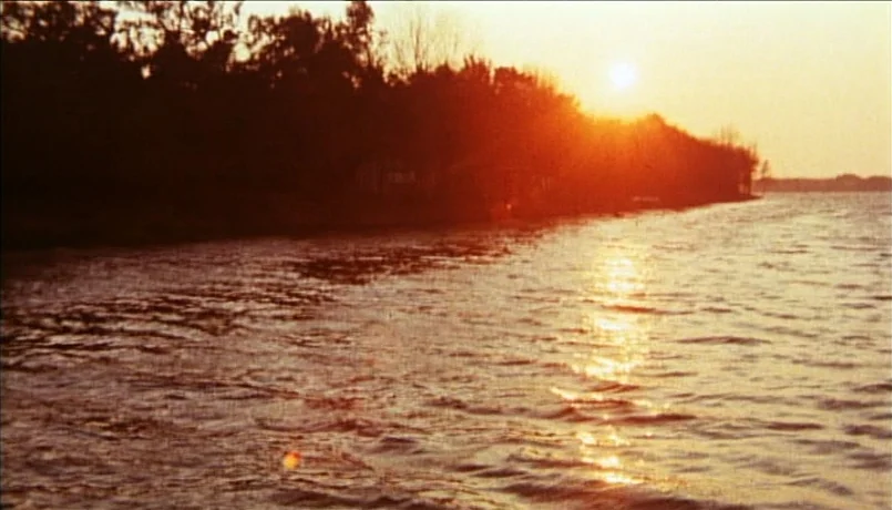 A Bay of Blood (1971) movie still: establishing — A calm lake reflects the low-hanging sun near a dense, silhouetted tree line. Rippling water…