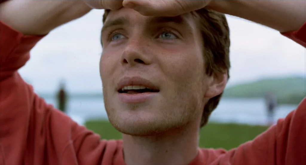 28 Days Later (2002) movie still: closeup — A man stands outdoors near a blurry body of water and green vegetation. He holds his hands above his…