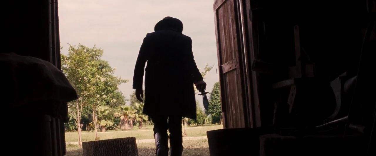 Django Unchained (2012) movie still: fullbody — A dark, wood-planked barn structure opens up to a bright, grassy outdoor field. A man wearing a long…