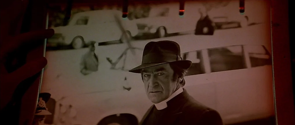 The Omen (1976) movie still: closeup — A close-up view of a photograph showing an older man in a clerical collar. The photograph is being held by a…