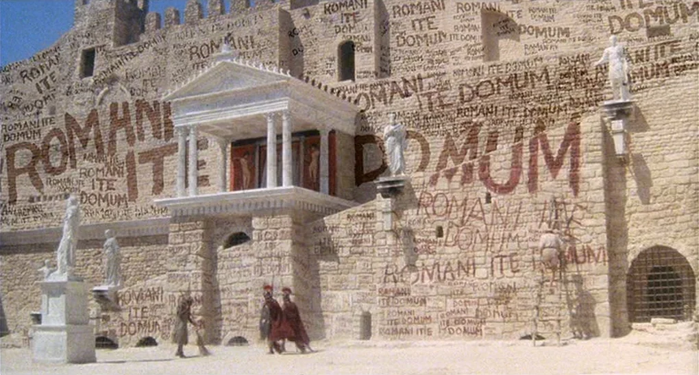 Life of Brian (1979) movie still: wide — A massive limestone wall of an ancient structure is covered in repetitive painted graffiti. A man on a…