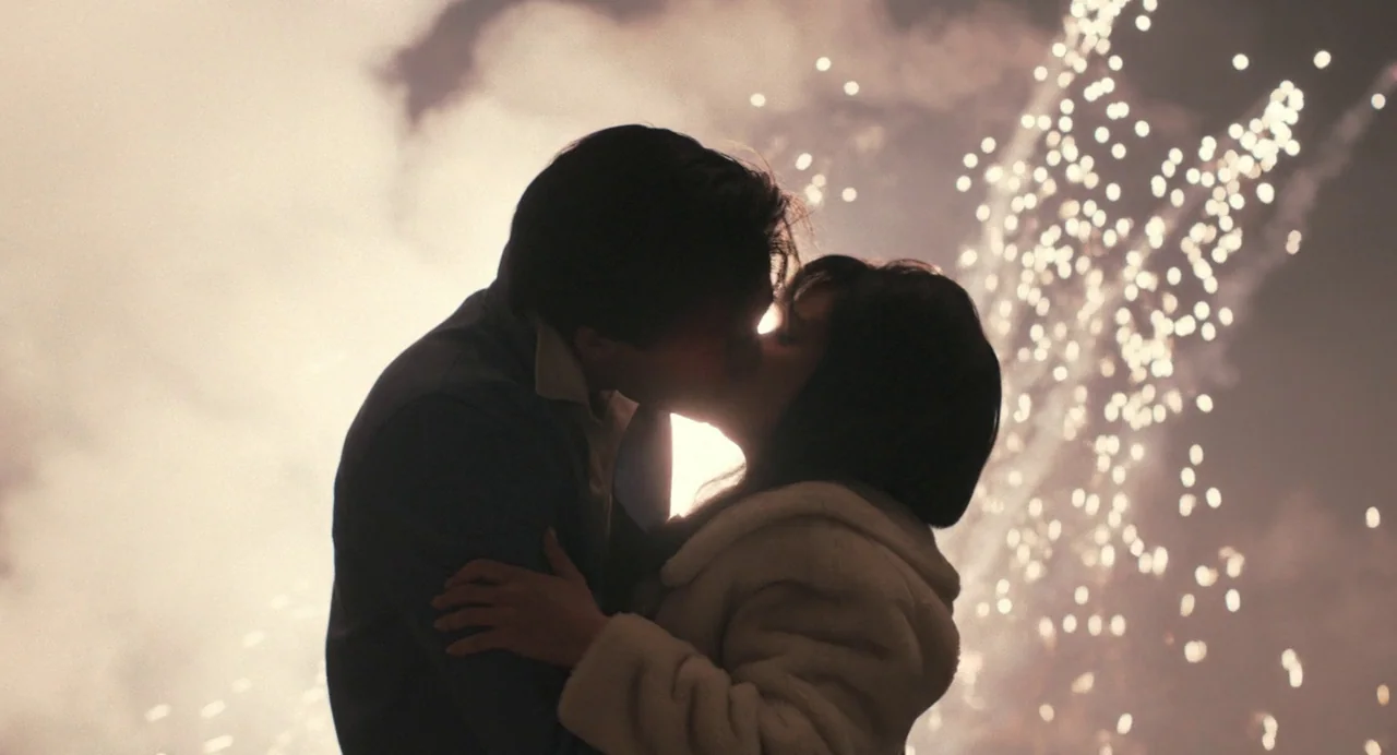 Priscilla (2023) movie still: medium — A night sky is filled with bright white light from large, exploding fireworks displays. Two people stand in a…