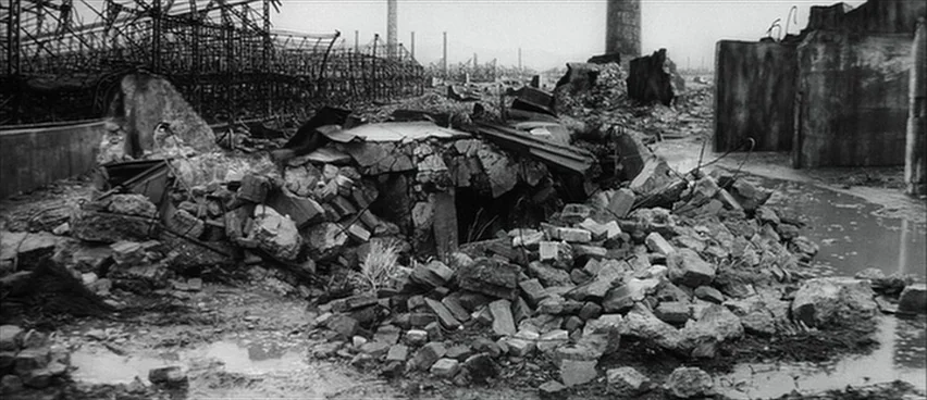The Bad Sleep Well (1960) movie still: establishing — A desolate field of concrete rubble and industrial debris under an overcast sky. Puddles of…