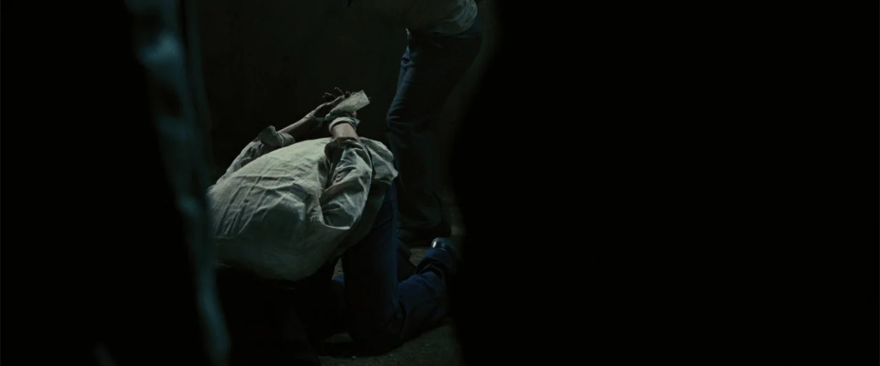 Argo (2012) movie still: medium — A dark, sparsely lit interior space with concrete walls and limited illumination. A person kneels on the floor…