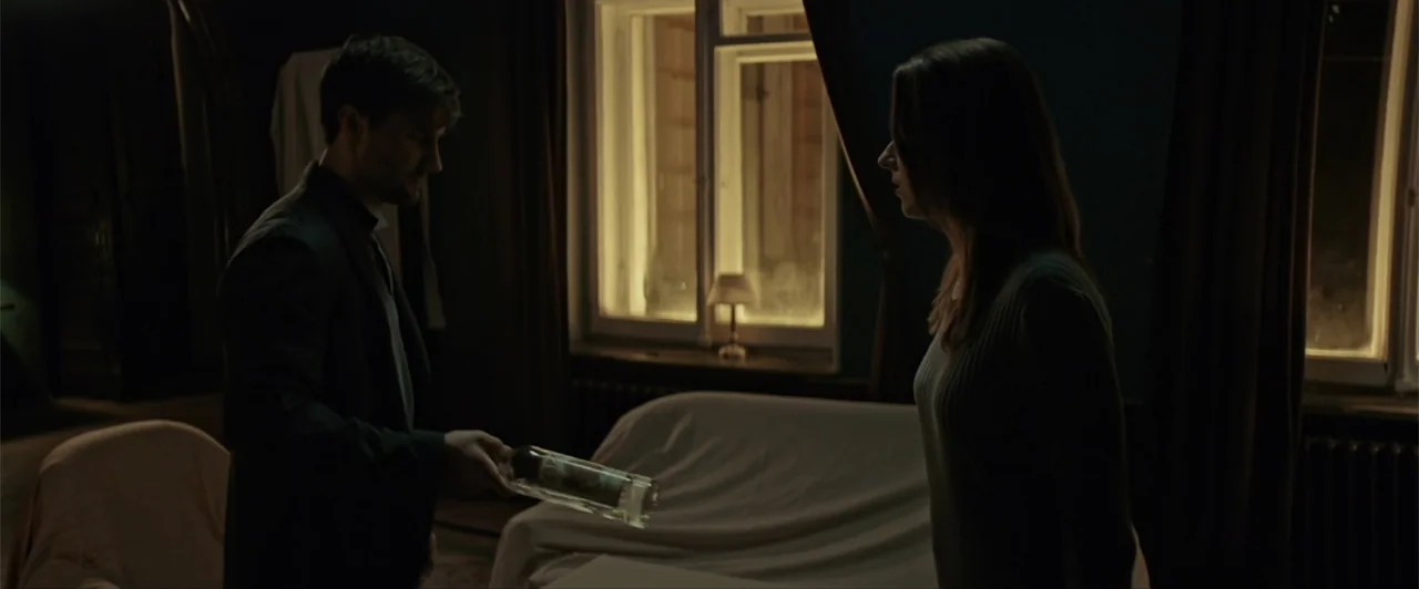 La herencia (2010) movie still: medium — A dimly lit room at night contains two people standing near furniture covered in sheets. A man offers a…