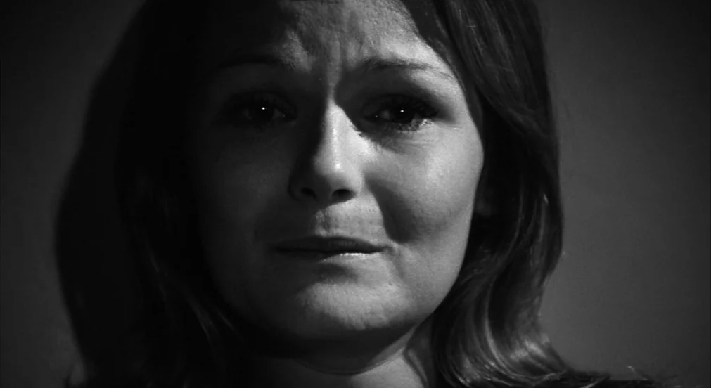 Lenny (1974) movie still: closeup — A dark, unadorned interior space frames a woman from the chest up. The woman stares directly into the camera…