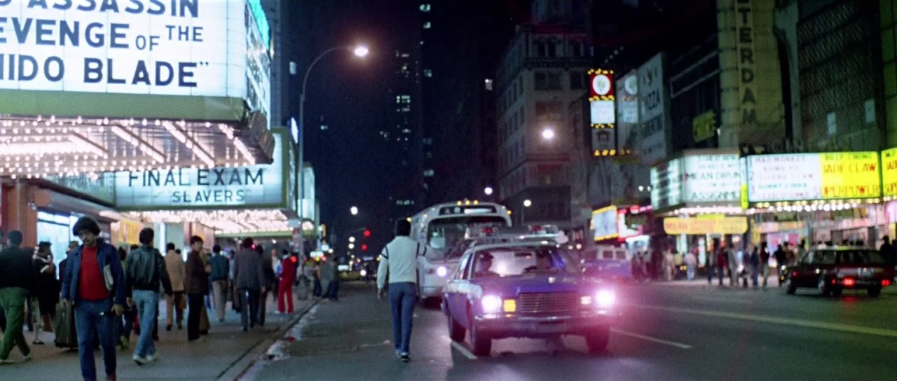 The New York Ripper (1982) movie still: wide — A bustling 1980s urban street at night is illuminated by bright movie theater marquees. Pedestrians…