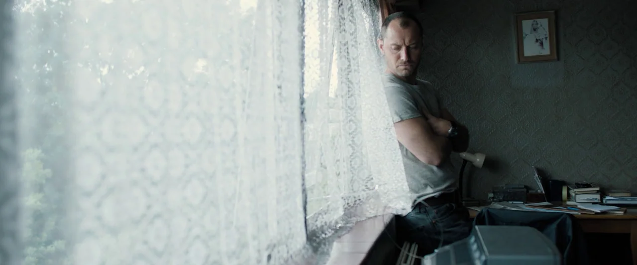 Black Sea (2014) movie still: medium — A man stands in a bedroom interior with textured wallpaper and lace curtains. He leans against the window…