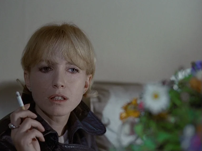 The Third Generation (1979) movie still: closeup — A woman sits indoors in front of a blurred bouquet of colorful flowers. She holds a lit cigarette…