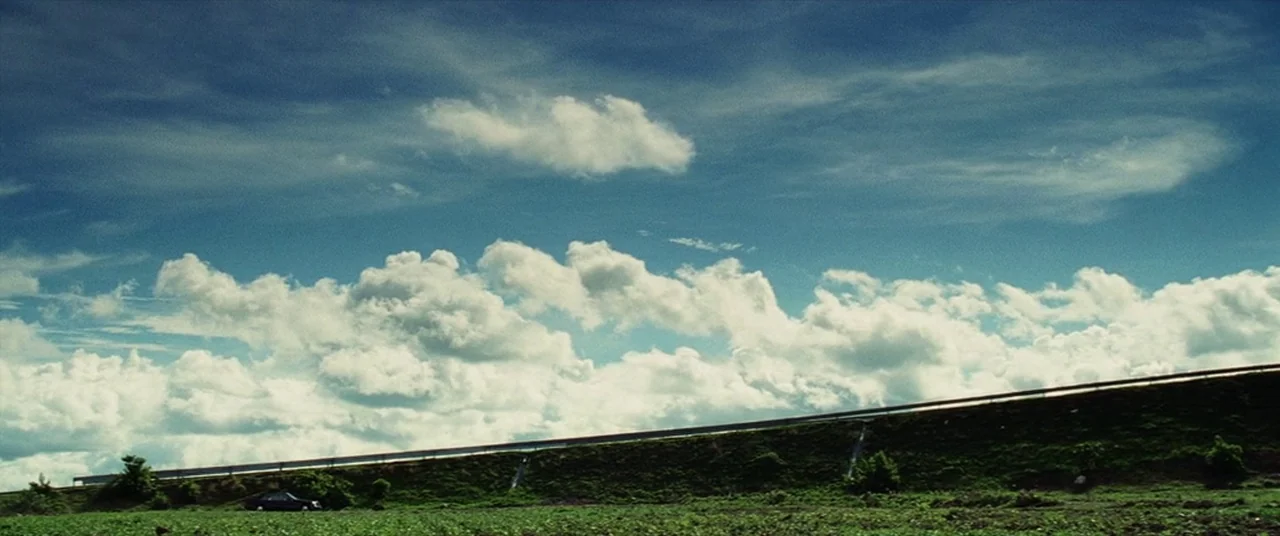 Man on Fire (2004) movie still: establishing — A vast blue sky filled with white cumulus clouds hangs over a green embankment. A single black car…