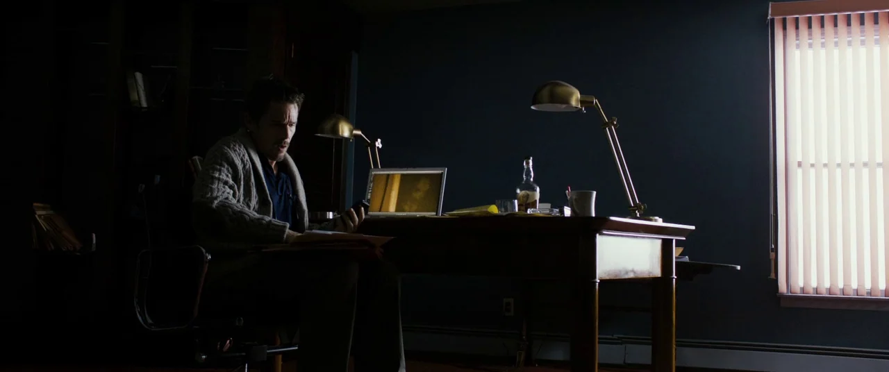 Sinister (2012) movie still: medium — A dark, dimly lit study with a wooden desk holding a laptop and lamp. A man sits at the desk, glancing at a…