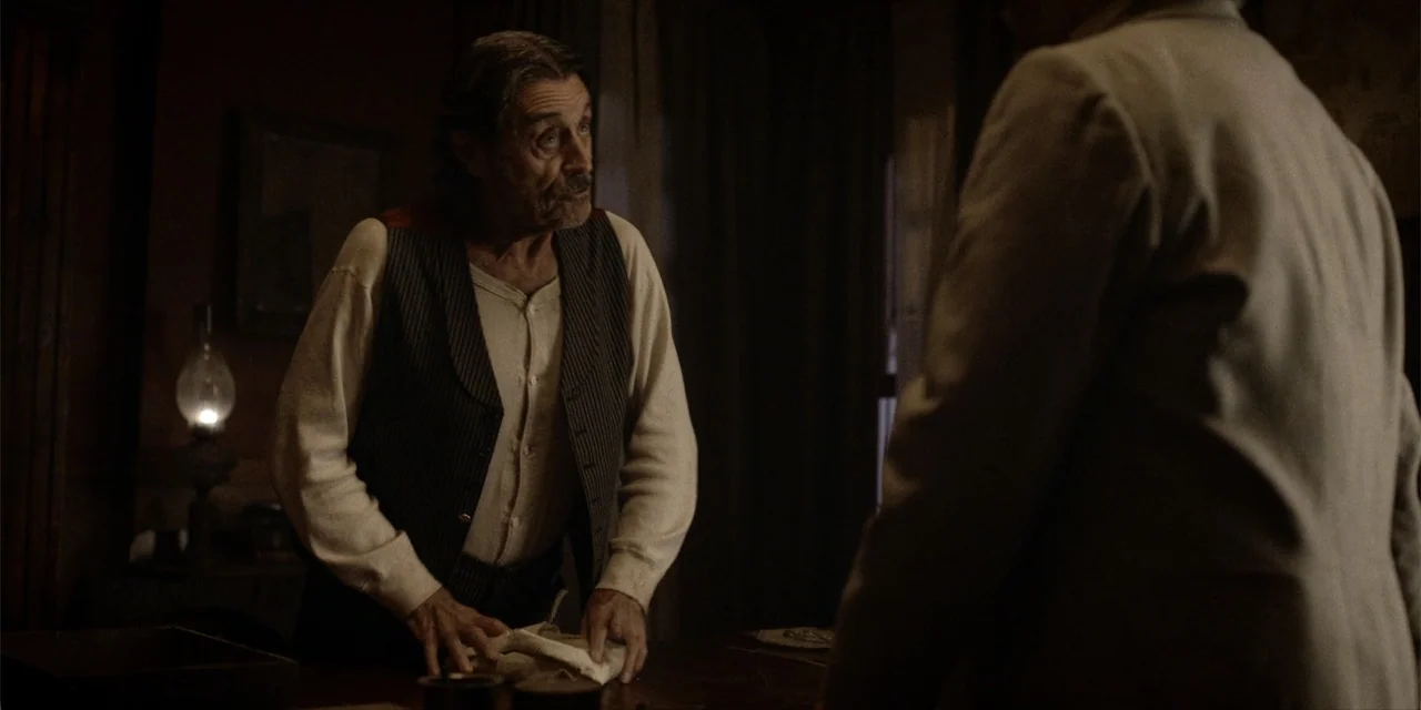 Deadwood: The Movie (2019) movie still: medium — A dimly lit 19th-century room features wood panels and a glowing oil lamp. An older man in a vest…