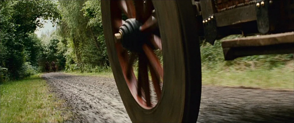 Your Highness (2011) movie still: closeup — A wooden carriage wheel rolls along a gravel and dirt path lined with trees. Two figures on horseback…