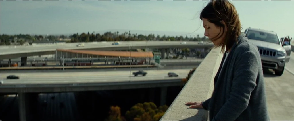 Cake (2014) movie still: medium — An elevated concrete highway overpass stretches over a multi-level traffic intersection in broad daylight. A woman…