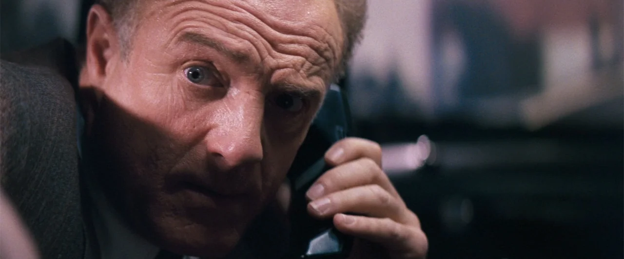 The Eraser (1977) movie still: closeup — An interior office space is dark with shadows obscuring the background. A mature man holds a black…